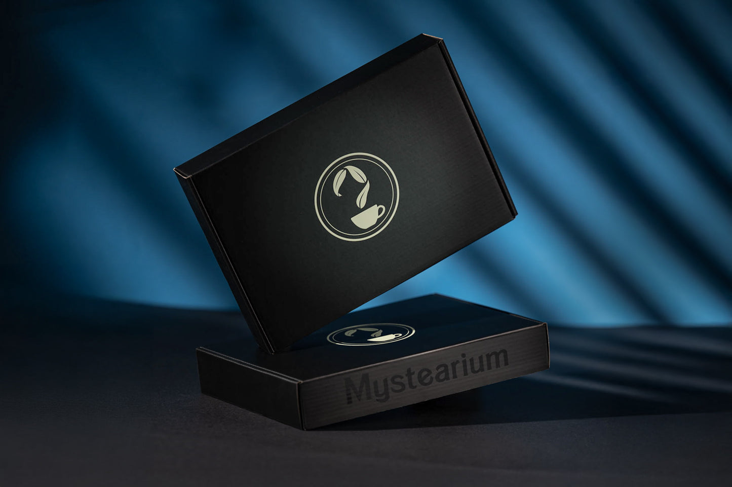 Two sleek black Mystearium gift boxes displayed against a blue background, one standing upright and the other lying flat, both featuring the Mystearium logo with a teacup and leaves, evoking a sense of elegance and premium tea branding.