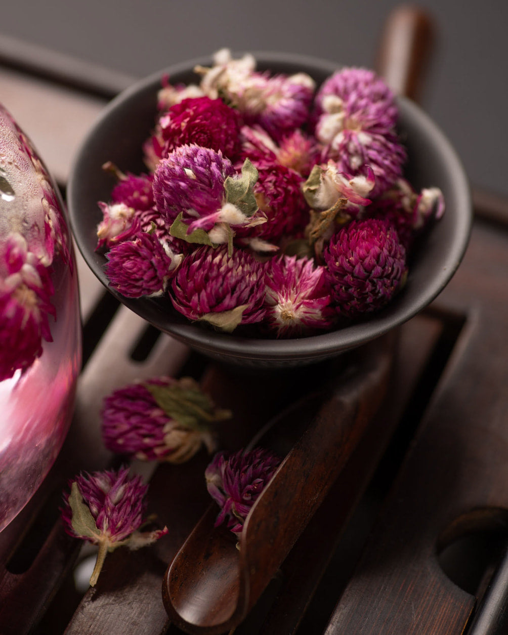 Eine Keramikschale, gefüllt mit leuchtend violetten getrockneten Kugelnamaranthblüten, steht auf einem traditionellen hölzernen Teetablett, daneben liegen verstreute Blüten und ein Glasgefäß ist teilweise zu sehen, in dem die Blüten in heißem Wasser ziehen.
