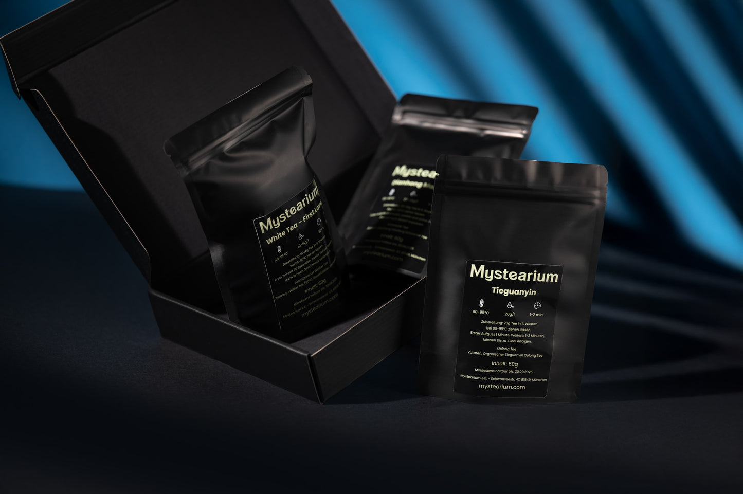 A premium Mystearium tea gift set displayed against a dark blue background, featuring three matte black resealable pouches labeled with tea names, with one pouch standing outside the sleek black box showcasing product details and brewing instructions.