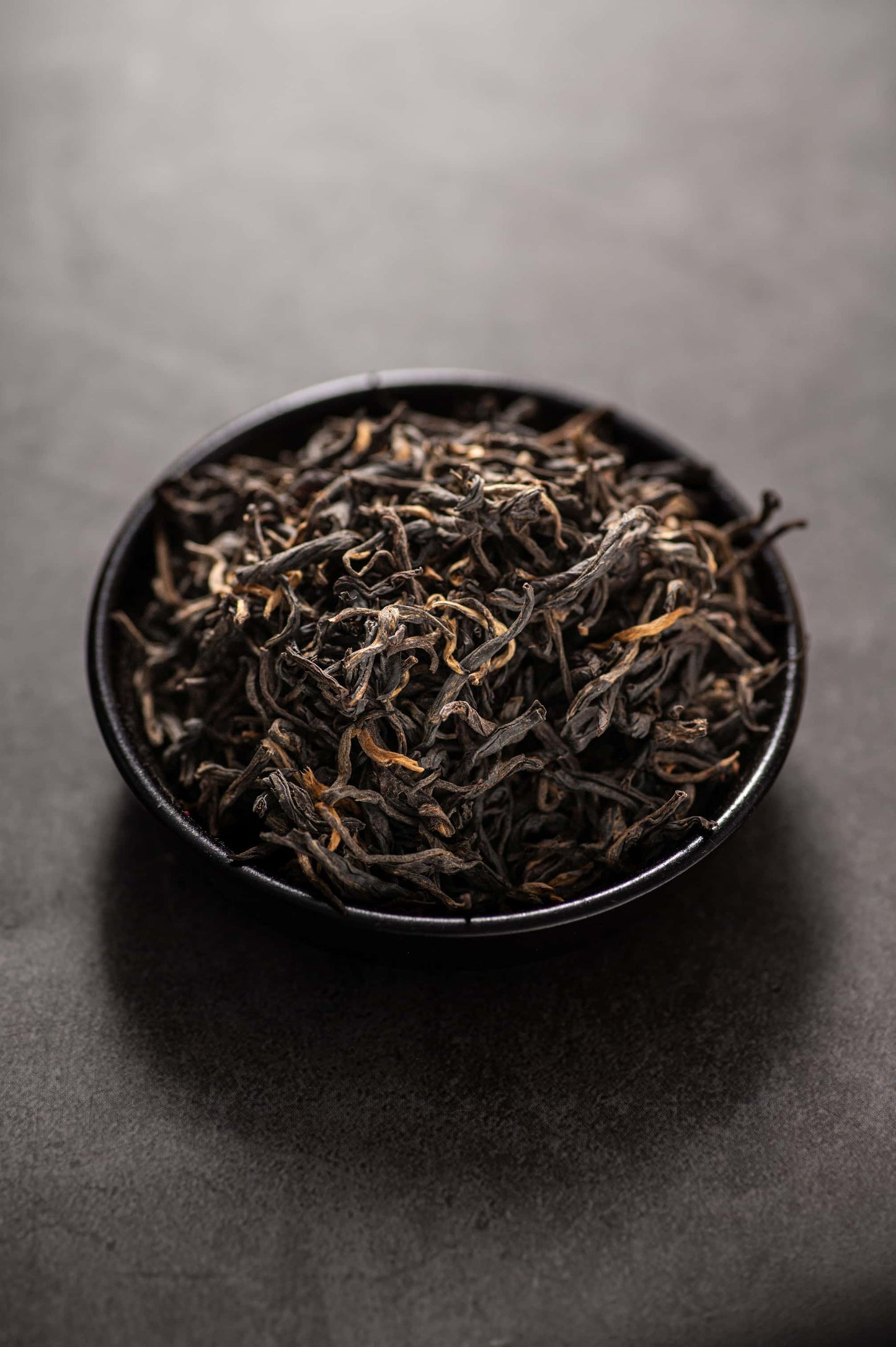 Eine Schale gefüllt mit hochwertigen getrockneten Dianhong Maofeng Schwarzteeblättern, die sich durch fest gedrehte Stränge mit goldenen Spitzen und einer glänzenden schwarzen Farbe auszeichnen. Der Tee ist bekannt für sein reichhaltiges Honig- und Fruchtaroma, seinen milden, süßen Geschmack und seine hochwertige traditionelle Verarbeitung, die den EU-Bio-Standards entspricht.