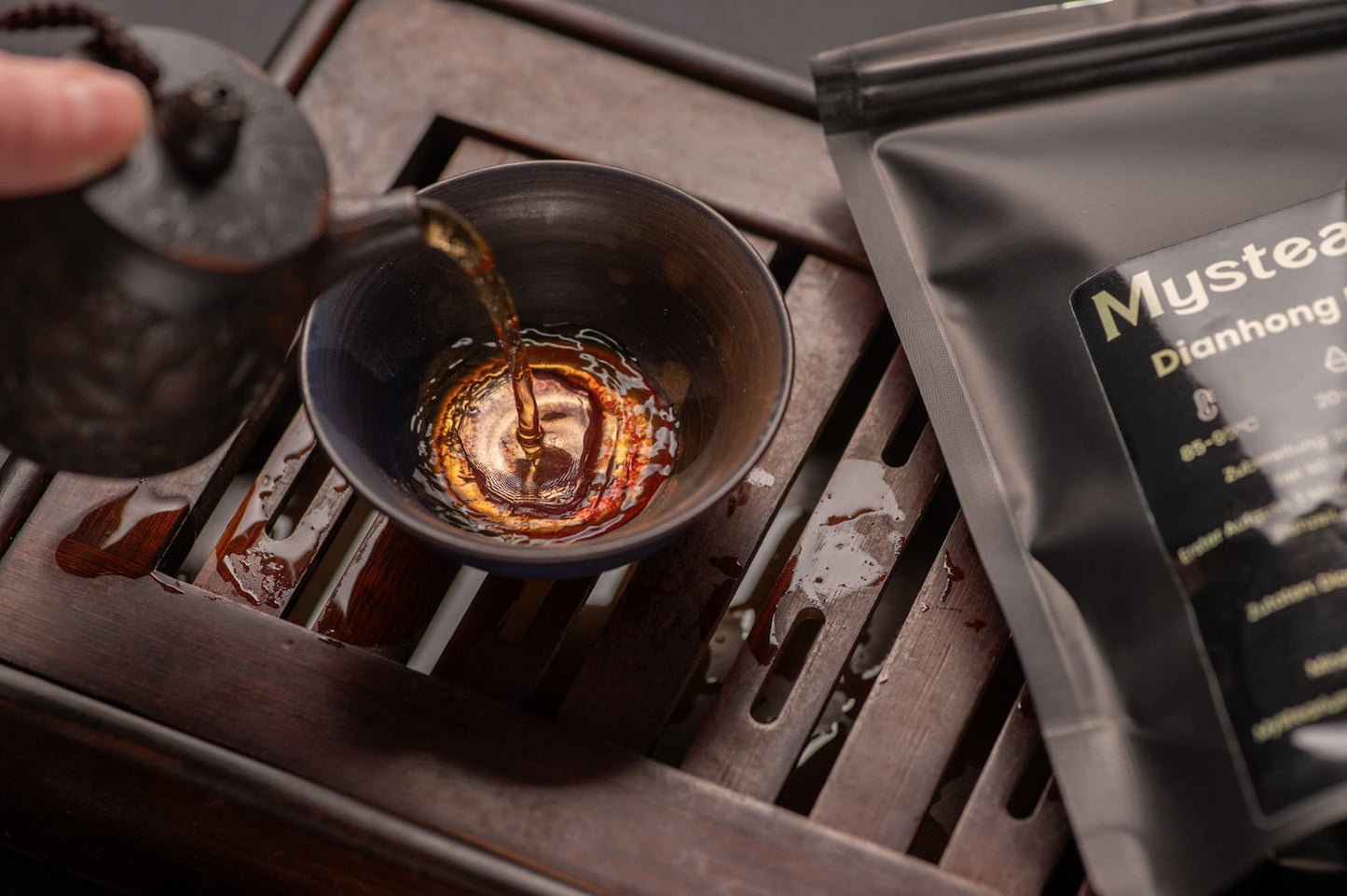 Person pouring tea into a black teacup with a Mystearium tea bag in the background.