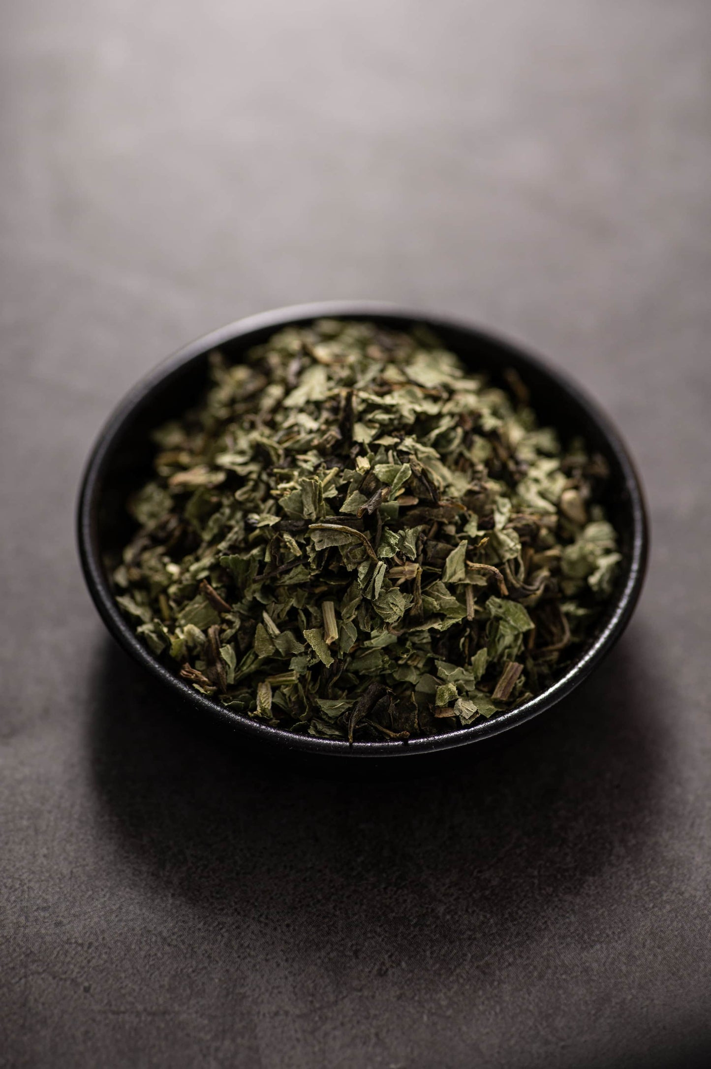A bowl filled with a dry green tea blend called ‘Green Tea Mint,’ made from finely cut green tea leaves and dried mint leaves.