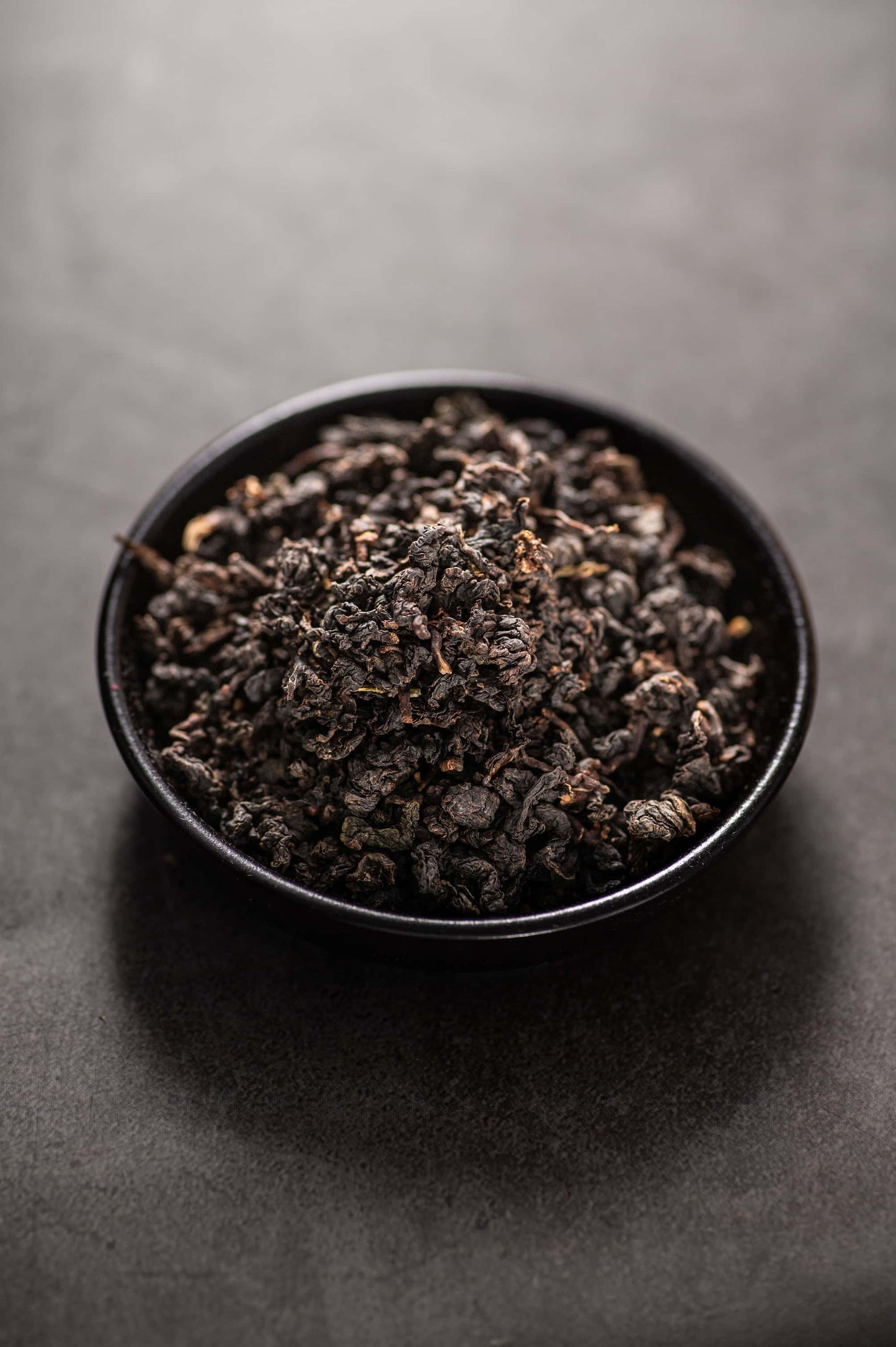 A black bowl filled with dry, tightly rolled Sechuong Oolong tea leaves, deep green to dark green in color with some reddish edges, known for its floral and fruity aroma, rich mellow flavor, and organically certified quality that meets EU standards.
