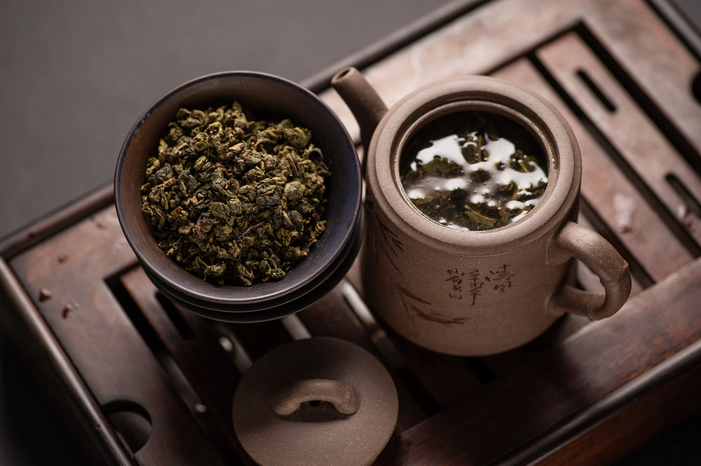 Ein traditionelles Teeservice bestehend aus einer rustikalen Ton-Teekanne mit aufgegossenem Tieguanyin-Oolong-Tee und einer dunklen Keramikschale mit getrockneten, gerollten Teeblättern, alles auf einem hölzernen Teetablett.