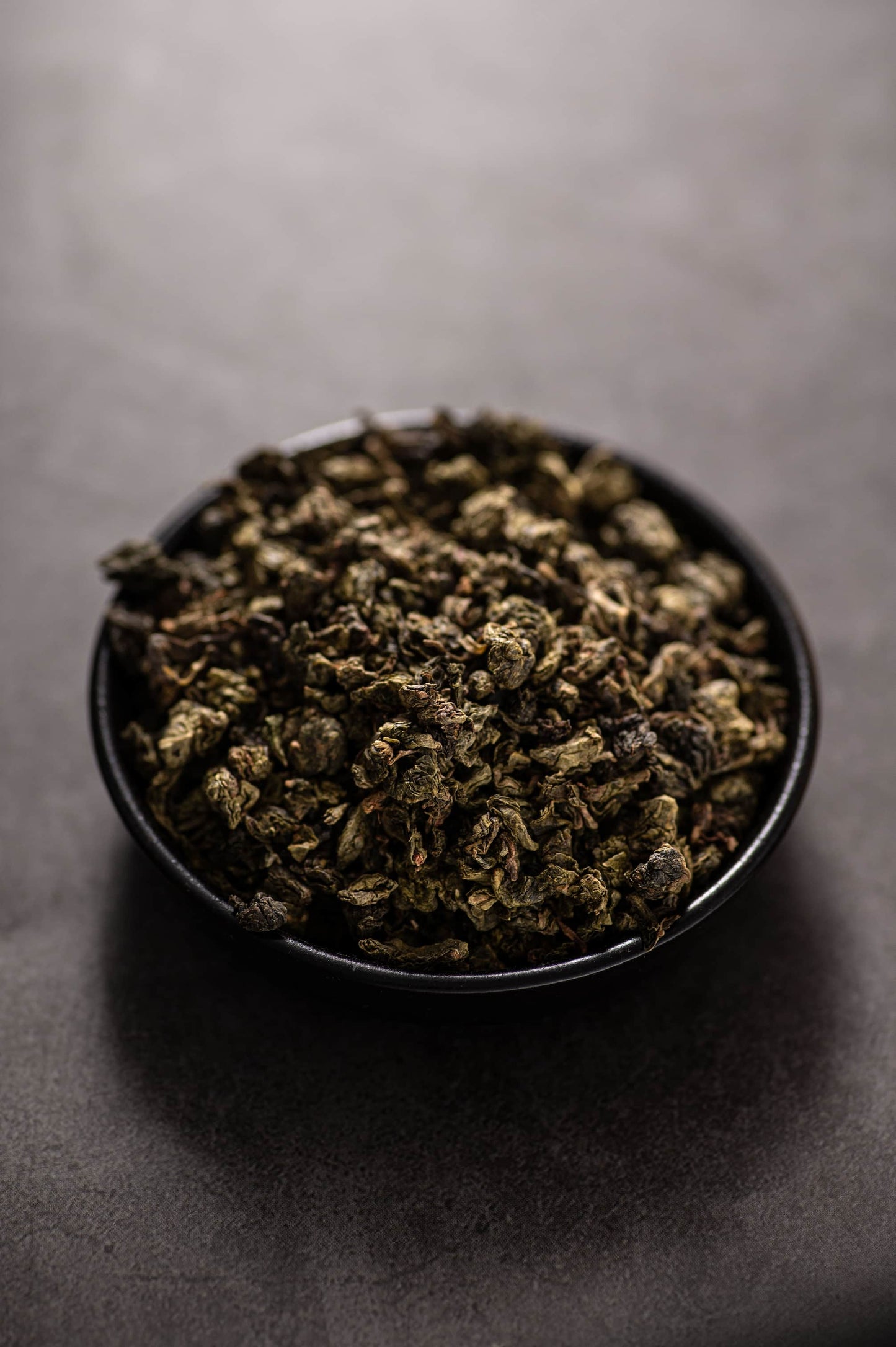 A black bowl filled with dry, rolled Organic Tieguanyin oolong tea leaves in deep green and brown tones, known for its intense orchid aroma, fresh and smooth taste, and organically grown free from pesticides and chemical fertilizers.
