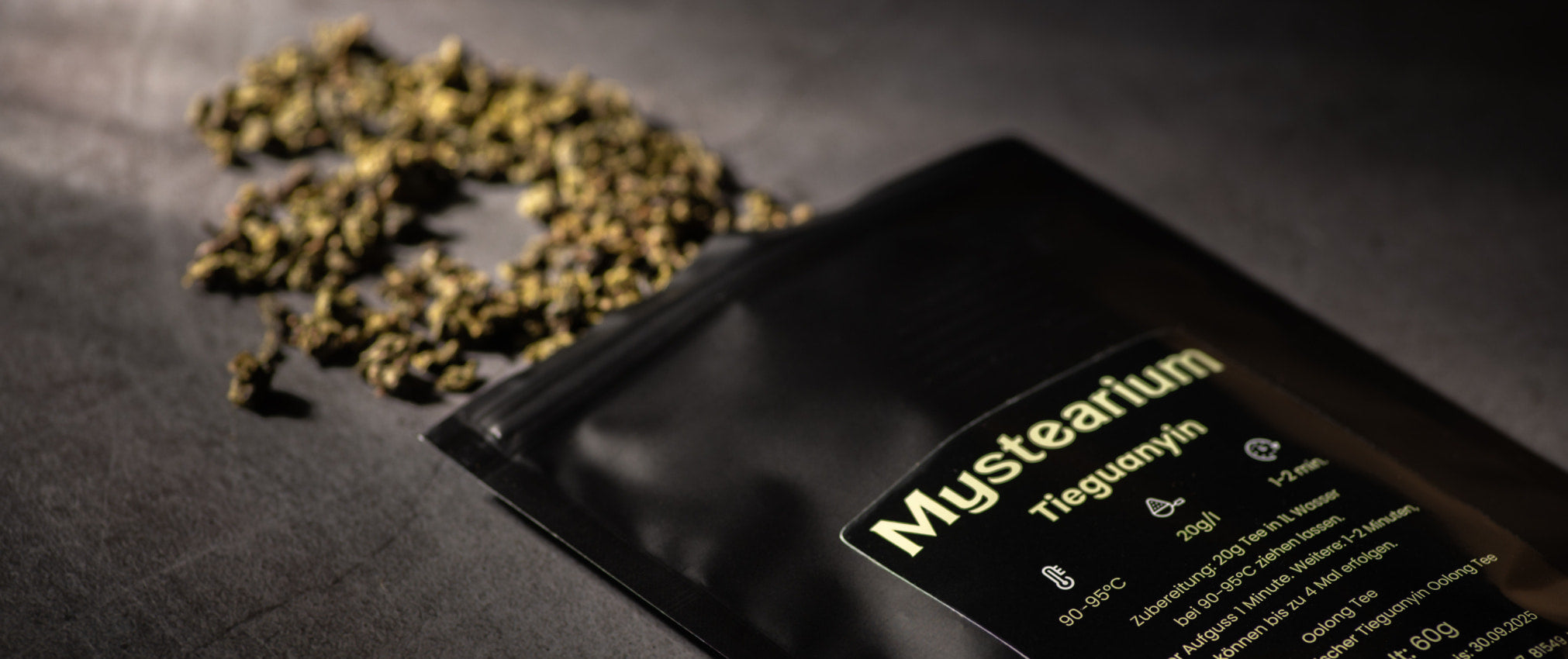 Nahaufnahme einer Verpackung des Oolong-Tees Tieguanyin der Marke Mystearium, die auf einer dunklen Oberfläche liegt, daneben liegen einige lose, trockene Tieguanyin-Teeblätter verstreut. Auf dem Etikett sind Zubereitungshinweise angegeben.