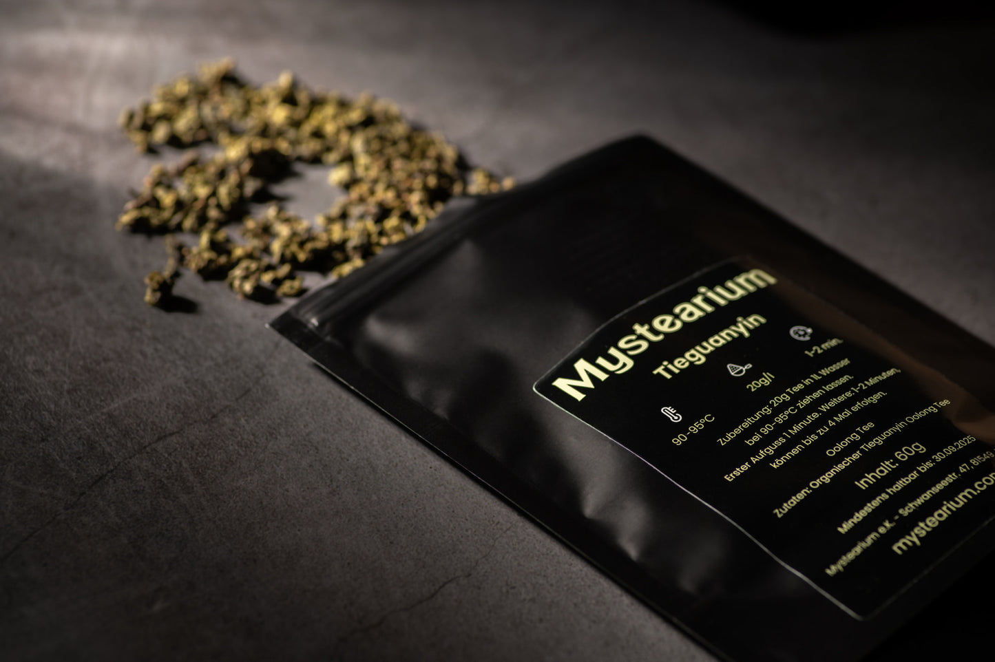 A close-up of Mystearium-branded Tieguanyin oolong tea packaging lying on a dark surface, with some loose, dry Tieguanyin tea leaves scattered beside it. The label shows brewing instructions.