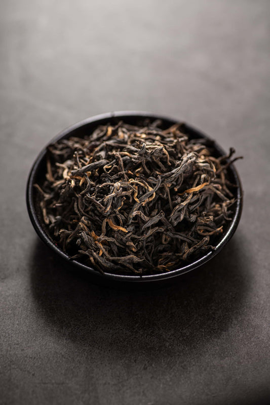 A bowl filled with premium dry Dianhong Maofeng black tea leaves, showing tightly twisted strands with golden tips and a lustrous black color, known for its rich honey and fruity aroma, mellow sweet taste, and high-quality traditional processing that meets EU organic standards.