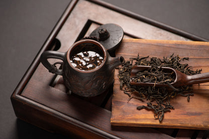 A traditional dark clay teapot filled with steeping Dianhong Maofeng black tea sits on a wooden tea tray beside a wooden scoop holding long, twisted dry black tea leaves, evoking a rustic and artisanal tea preparation setting.