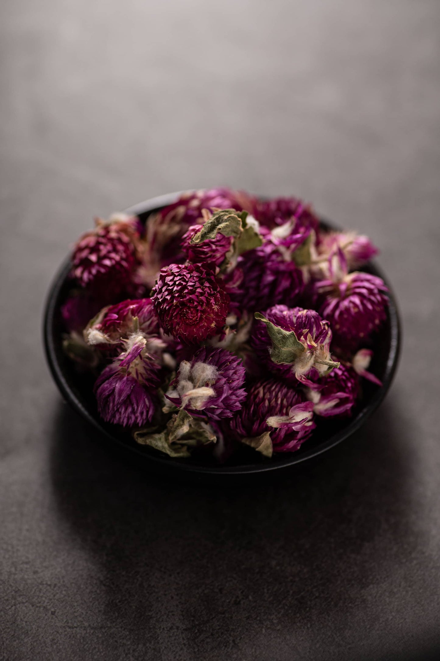 A black bowl filled with dried purple globe amaranth flowers, showcasing their vibrant magenta color and textured petals, prepared for tea blending or infusion.