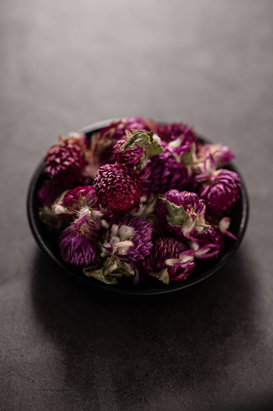 A black bowl filled with dried purple globe amaranth flowers, showcasing their vibrant magenta color and textured petals, prepared for tea blending or infusion.