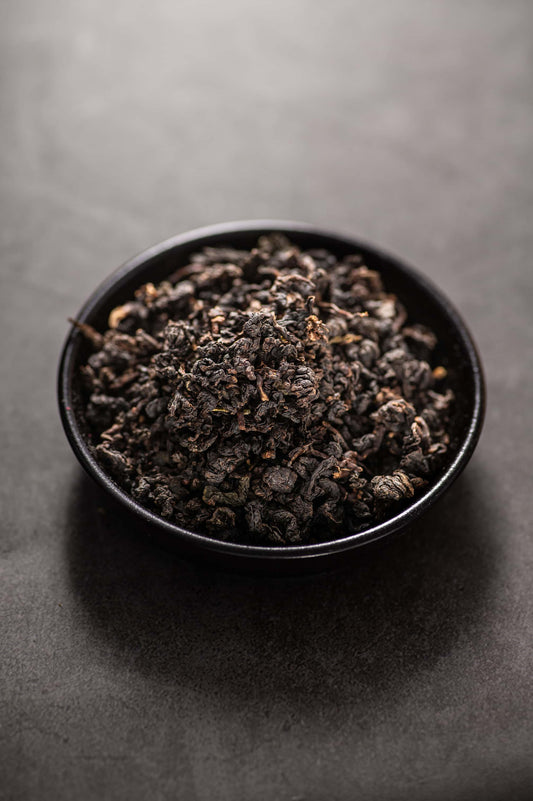 A black bowl filled with dry, tightly rolled Sechuong Oolong tea leaves, deep green to dark green in color with some reddish edges, known for its floral and fruity aroma, rich mellow flavor, and organically certified quality that meets EU standards.