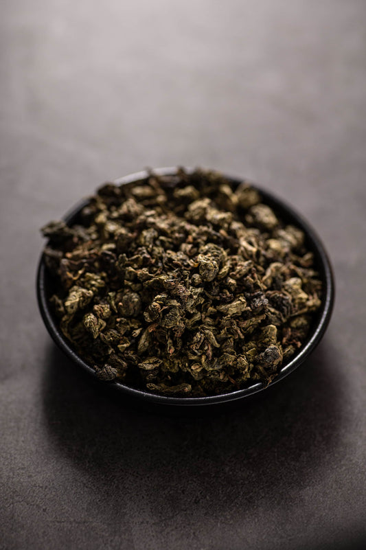 A black bowl filled with dry, rolled Organic Tieguanyin oolong tea leaves in deep green and brown tones, known for its intense orchid aroma, fresh and smooth taste, and organically grown free from pesticides and chemical fertilizers.