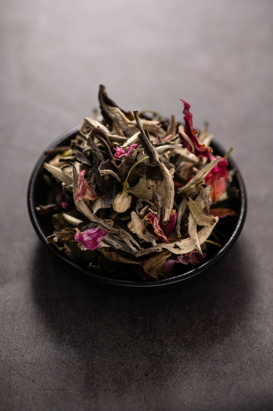 A bowl of dry loose leaf white tea blend called ‘White Tea First Love,’ featuring delicate silver-tipped white tea leaves (90%) mixed with dried rose petals and jasmine buds.
